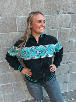 Woman wearing a black jacket with teal and white pattern standing against a brick wall.