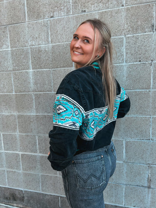Woman wearing a black sweater with turquoise patterns against a gray brick wall.