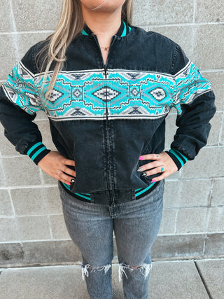 Person wearing a black denim jacket with teal and white patterned accents, standing against a stone wall.