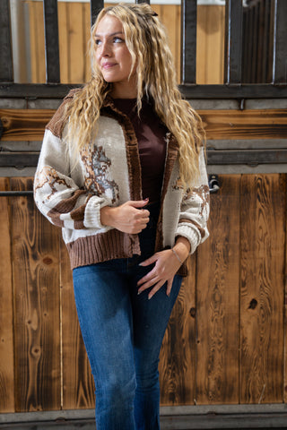 Woman wearing a patterned cardigan over a dark top and blue jeans, standing against a wooden wall.