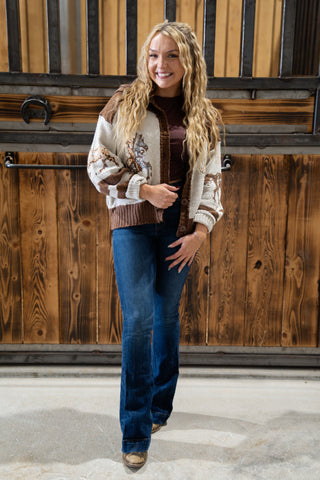Woman wearing a patterned sweater and blue jeans standing in front of a wooden wall.