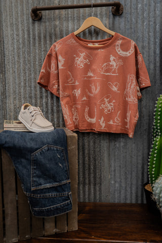 Vintage-style t-shirt with graphic design on a hanger, paired with jeans and sneakers against a corrugated metal background.