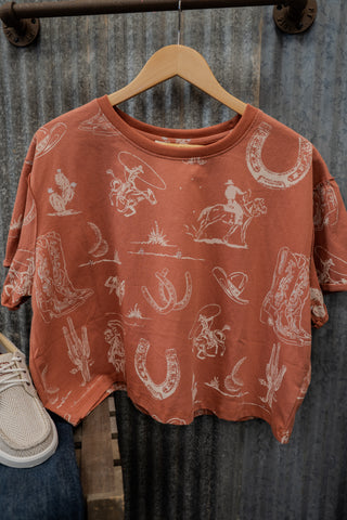 Red t-shirt with white graphic designs on a wooden hanger against a rustic metal wall.
