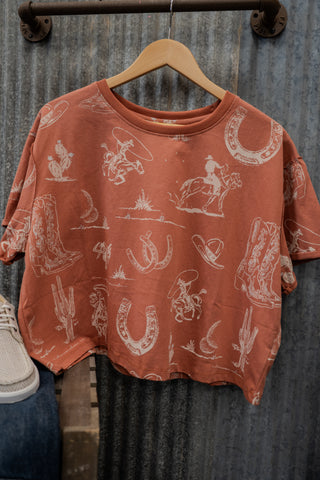 Rust-colored t-shirt with white graphics on a wooden hanger against a rustic metal wall.