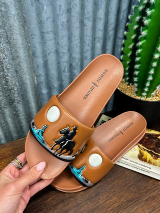 Brown slide sandals with decorative elements held by a hand on a wooden surface.