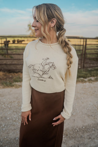 Woman wearing a cream sweater with a horse graphic and brown skirt in a rural setting.