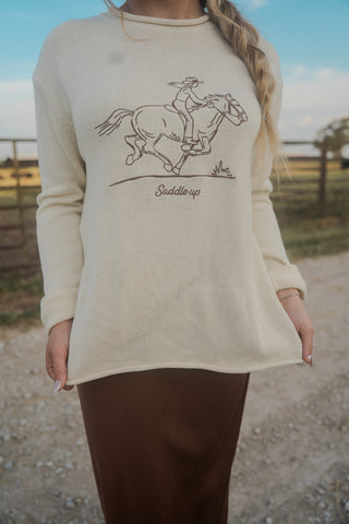 Person wearing a cream sweater with a horse and rider graphic and 'Saddle up' text, standing outdoors.