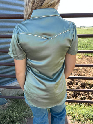 Person wearing a green shirt with white piping standing in front of a metal fence and grassy field.