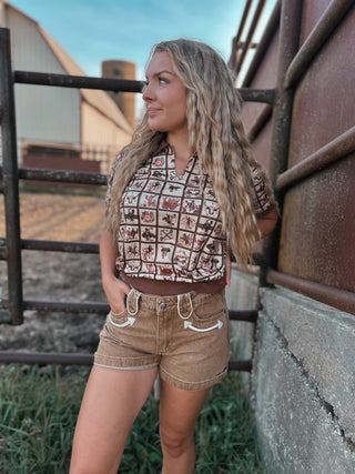 Woman in a checkered top and shorts standing next to a rusted metal wall with a barn in the background.
