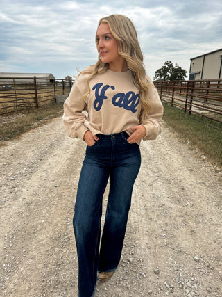 Woman wearing a beige sweatshirt with 'Y'all' text and blue jeans on a dirt road.