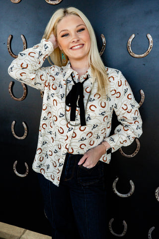 Woman wearing a patterned blouse with a black bow tie in front of a dark background with horseshoe decorations.