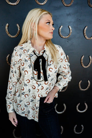 Woman wearing a patterned blouse with a black bow tie against a dark background with horseshoe decorations.