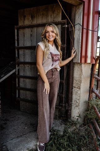 Woman standing in a rustic setting wearing a sleeveless top and high-waisted pants.