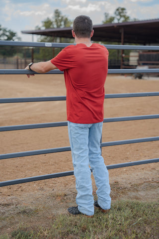 Man in a red shirt and light blue jeans standing by a metal fence in an outdoor setting.