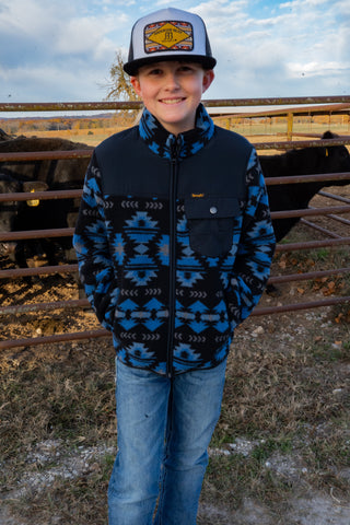Child wearing a patterned jacket and cap standing in an outdoor setting with a fence and sky in the background.