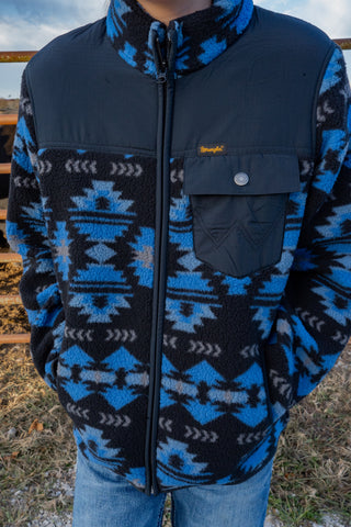 Person wearing a blue and black patterned jacket with a brand logo, standing outdoors.