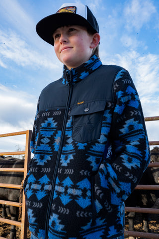 Person wearing a blue and black patterned jacket with a cap, standing outdoors with a cloudy sky.