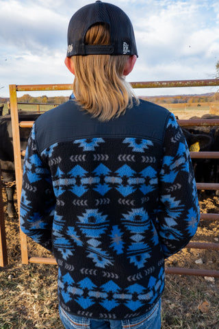 Person wearing a blue patterned jacket and black cap in an outdoor setting with a fence and sky.