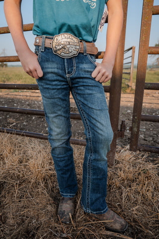 Person wearing blue jeans and a green shirt with a belt buckle, standing in front of a metal gate.