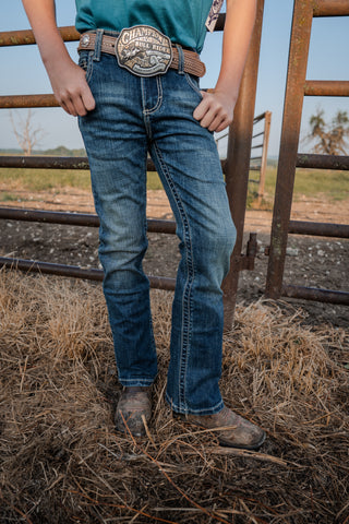 Person wearing blue jeans and a belt with a prominent brand logo, standing in front of a metal gate.