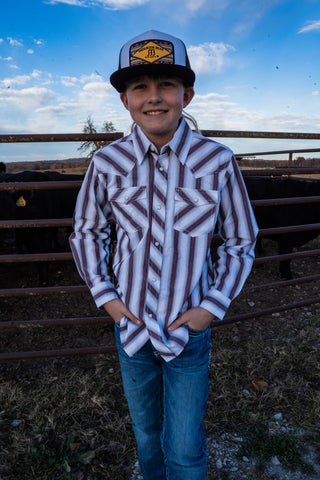 Child wearing a patterned shirt and cap standing in an outdoor setting with a fence and sky in the background.
