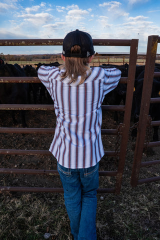 Person wearing a striped shirt and cap, standing behind a metal fence looking at cows in a field.