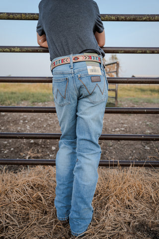 Person wearing blue jeans and a patterned belt standing in a field with a metal fence in the background.