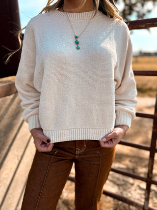 Person wearing a beige sweater and brown pants with a blurred outdoor background