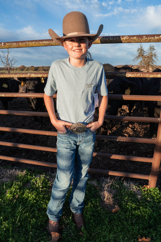 Person wearing a cowboy hat and standing in front of a rustic fence with cattle in the background.