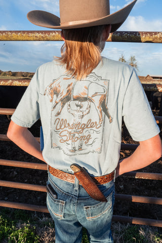 Person wearing a light gray 'Wrangler Strong' t-shirt with a cowboy hat and belt, standing in front of a metal fence.