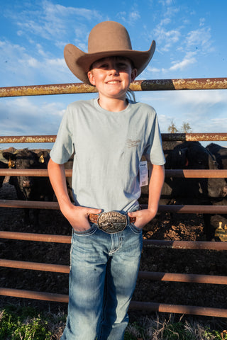Person wearing a cowboy hat and belt buckle standing in front of a metal fence with cows in the background.