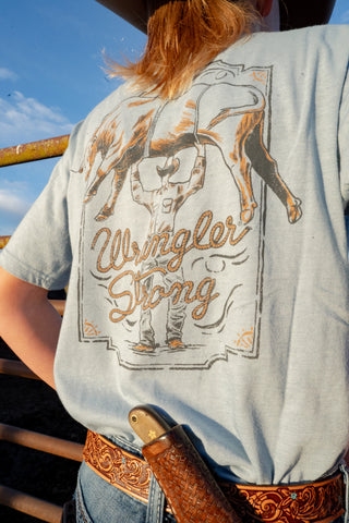 Person wearing a 'Wrangler Strong' t-shirt with a graphic design on the back, outdoors.